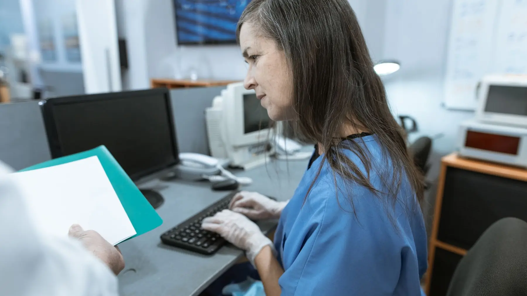 Recepção de uma clínica no meio do expediente, atendente registrando no computador enquanto recebe uma pasta de documentos